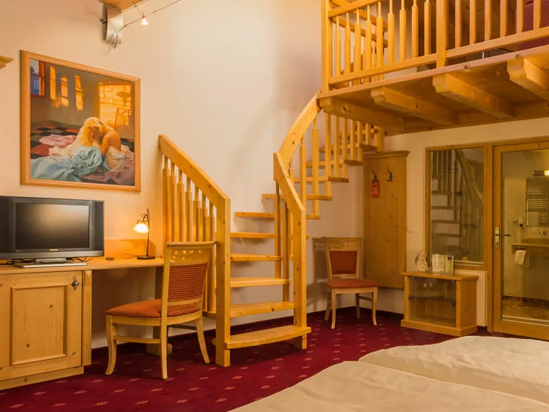 Zimmer mit Doppelbett, offener Galerie-Ebene über der Holztreppe und verglastem Badezimmer